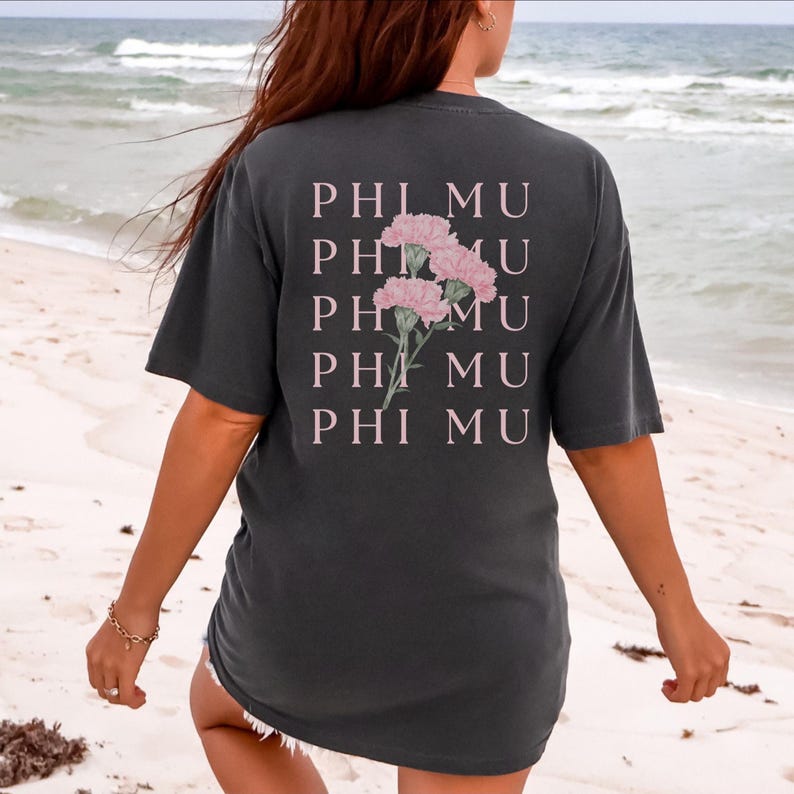 Op de afbeelding: Donkergrijs t-shirt met de tekst "PHI MU" en een roze bloemenontwerp. Het casual t-shirt is geschikt voor dagelijks gebruik. De foto is genomen op een strand.