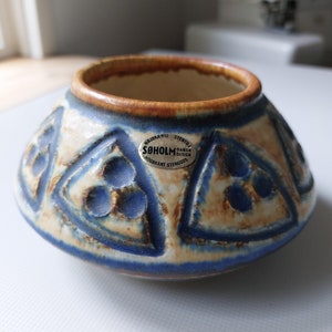 Peut inclure: Un petit pot en céramique bleu et blanc avec un bord brun. Le pot a un motif géométrique avec des triangles et des cercles. Le pot porte une étiquette qui indique "Soholm Dansk Design Højbrændt Stengods".