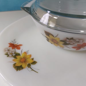 Könnte beinhalten: Eine weiße Auflaufform mit Glasdeckel und passendem Teller. Die Auflaufform und der Teller sind mit einem Blumenmuster mit gelben, orangefarbenen und weißen Blumen mit grünen Blättern verziert.
