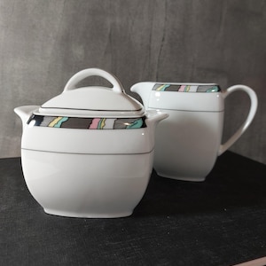 Puede incluir: Un azucarero de porcelana blanca con tapa y una jarra de leche a juego. Ambas piezas presentan una banda horizontal decorada con patrones geométricos en negro, gris, rosa y azul claro, junto con finas líneas negras. El azucarero tiene dos asas.
