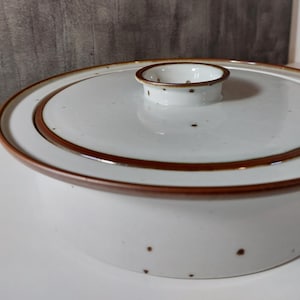 Könnte beinhalten: Eine weiße Keramik-Auflaufform mit Deckel. Die Schüssel hat einen braunen Rand und einen kleinen Griff auf dem Deckel. Die Schüssel hat kleine braune Sprenkel. Der Deckel hat einen braunen Rand und einen kleinen Griff.