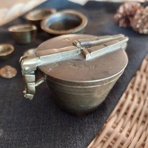 May include: A vintage brass container with a hinged lid and a decorative handle. The container has a worn, aged finish and is decorated with a series of raised lines.