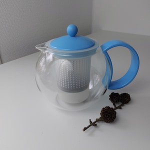 May include: A clear glass teapot with a blue lid and handle. The teapot has a built-in strainer for loose leaf tea.