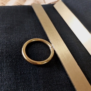 Könnte beinhalten: Ein goldener Metallring, der an einer Messingstange befestigt ist. Der Ring ist rund und hat eine glatte Oberfläche. Die Stange ist flach und hat eine gebürstete Oberfläche.