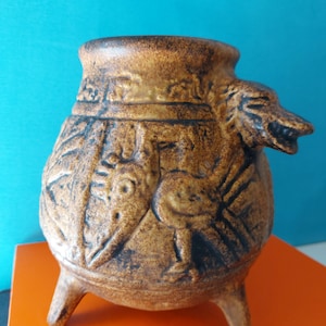 Op de afbeelding: Een bruine keramische pot met een wijde opening en drie korte pootjes. De buitenkant heeft een textuur met reliëfsculpturen van een vogel en geometrische patronen, aangevuld met een uitstekende dierenkop aan de zijkant van het object.