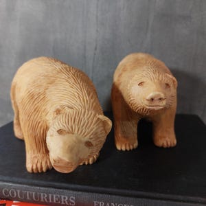 Puede incluir: Dos figuras de osos de madera tallada. Un oso está a cuatro patas, el otro está de pie. Los osos son de color marrón claro con tallas detalladas, mostrando la textura del pelaje. Se exhiben sobre una superficie oscura contra un fondo gris.