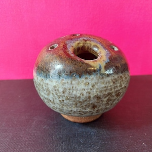 May include: A small, round, brown and white ceramic vase with a hole in the centre and five smaller holes around the top. The vase has a speckled glaze.