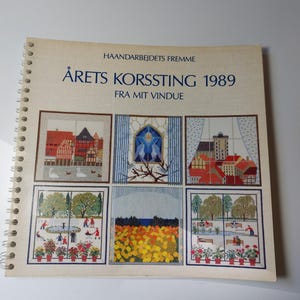 May include: A spiral-bound craft book titled HAANDARBEJDETS FREMME ÅRETS KORSSTING 1989 FRA MIT VINDUE. The cover features six distinct cross-stitch patterns depicting various outdoor scenes, including city buildings, a fountain, and flower fields.