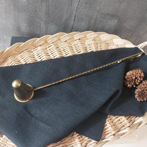 May include: A gold candle snuffer with a long, twisted handle. The snuffer is on a black cloth and a wicker basket.