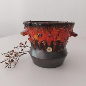 May include: A vintage ceramic planter with a drip glaze in shades of orange, yellow, and brown. The planter has two small handles and a gold sticker with the text "Manzanillo" on the side.