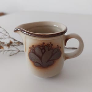 May include: A small, brown and beige ceramic creamer with a floral design. The creamer has a brown handle and a brown rim.