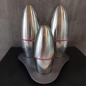 Puede incluir: Un juego de tres saleros y pimenteros de plata en forma de huevo con detalles rojos. Los saleros están sobre una base triangular gris.