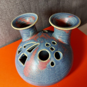 May include: A blue ceramic vase with two openings and decorative cutouts. The vase has a round body with a blue glaze and red accents. The cutouts include geometric shapes and circular holes. The vase is a unique decorative item.