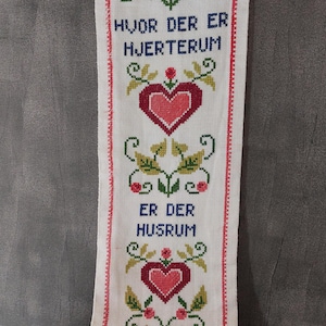 Könnte beinhalten: Bestickter Wandbehang mit dem Text "HVOR DER ER HJERTERUM ER DER HUSRUM". Das Design zeigt Herzen, Blumen und Blätter in Rot, Grün und Blau auf weißem Hintergrund, umrahmt von einem roten Rand.