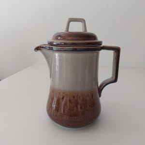 Puede incluir: Una jarra de crema de cerámica marrón y blanca con tapa y asa. La jarra tiene un esmalte marrón con un esmalte blanco en la mitad superior. La tapa tiene un esmalte marrón con un esmalte blanco en la parte superior.