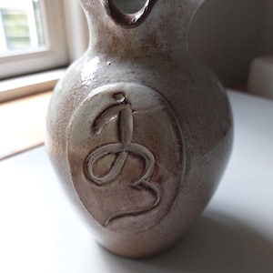 Peut inclure: Pichet en céramique fait à la main, de ton terreux et moucheté. Le pichet présente un motif en relief unique sur le devant. Le bord a une forme distinctive, et le design général suggère une esthétique rustique et artisanale.