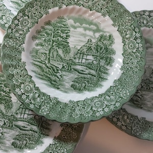 Może przedstawiać: Kilka zielono-białych ceramicznych talerzy z kwiatowym obramowaniem i sielankową sceną pośrodku. Talerze ułożone są w stos, prezentując szczegółowy wzór naczyń. Talerze są prawdopodobnie przeznaczone do serwowania potraw.