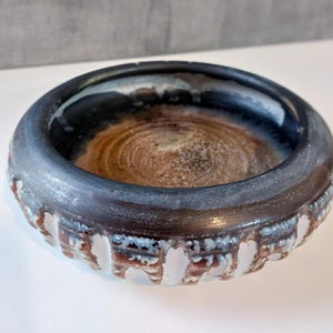 Può includere: Una ciotola in ceramica fatta a mano con un bordo blu scuro e un interno strutturato e terroso. L'esterno della ciotola presenta un motivo decorativo di elementi verticali bianchi e marroni. Un pezzo di ceramica unico.