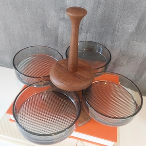 Puede incluir: Un soporte de servicio de madera con cuatro cuencos de vidrio transparente. El soporte tiene una base redonda y un mango alto y cilíndrico. Los cuencos tienen un patrón texturizado en forma de cuadrícula en el fondo.