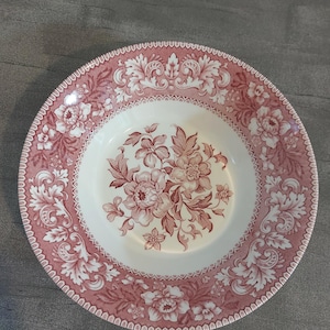 May include: A round ceramic bowl featuring a cream-coloured centre with a detailed pink floral illustration. The wide rim is decorated with a repeating pink and white pattern of intricate scrolling acanthus leaves and blooming flowers.