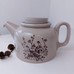 Peut inclure: Une théière en céramique beige avec un couvercle brun et un motif brun de deux oiseaux et de feuillage.