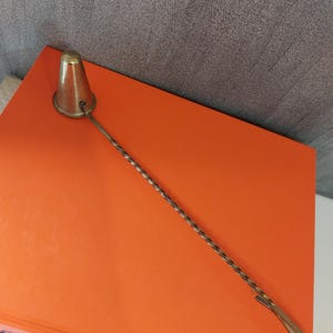 May include: A vintage brass cocktail muddler with a conical head and a twisted handle. The muddler is resting on an orange surface, with a gray background. The handle has a loop at the end.