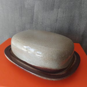 Puede incluir: Un recipiente para mantequilla de cerámica con un borde marrón y una tapa beige claro. La tapa tiene una superficie texturizada.