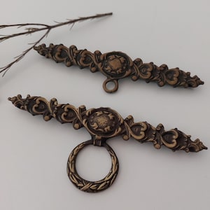 Könnte beinhalten: Zwei antike Schubladengriffe in Bronzefarbe mit aufwendigen floralen Designs. Einer hat einen Ringgriff, der andere eine kleine kreisförmige Öffnung. Die Griffe sind vor einem weißen Hintergrund mit einem Zweig einer Pflanze platziert.