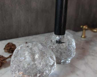 May include: Two clear, textured glass candle holders with a black candle. The candle holders have a spherical shape with a crackled ice-like appearance. The candle is inserted into one of the holders. The background is a marble surface.