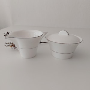 May include: White creamer and sugar bowl set with a silver trim. The creamer has a curved spout and the sugar bowl has a lid.