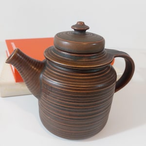 Può includere: Una teiera in ceramica marrone con un coperchio e un design a coste. La teiera ha un beccuccio e un manico.