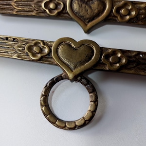 May include: Two antique brass door handles with a heart and flower design. Each handle has a ring pull and is decorated with a heart and two flowers.