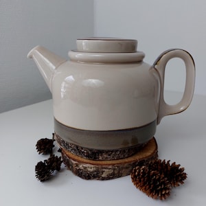 May include: A cream-colored ceramic teapot with a brown lid and a brown band around the bottom. The teapot is sitting on a wooden coaster.
