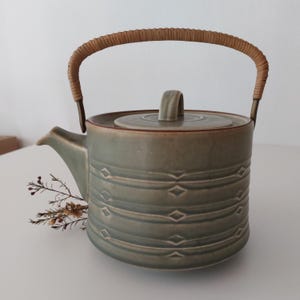 Puede incluir: Una tetera de cerámica verde con un asa trenzada y un patrón geométrico. La tetera tiene una tapa y un pico.