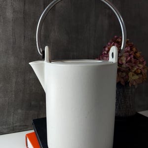 Puede incluir: Una tetera de cerámica blanca con un asa plateada. La tetera tiene un cuerpo cilíndrico y un pico. El asa se arquea sobre la parte superior. La tetera está colocada encima de un cuaderno negro y un cuaderno naranja.