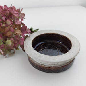 May include: A small, round ceramic dish with a white rim and a brown interior. The dish has a speckled glaze and a slightly raised lip.