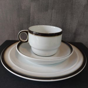 Op de afbeelding: Een witte keramische theekop en schotelset met een zwarte rand. De kop en schotel zijn beide effen wit met een enkele zwarte band rond de rand.
