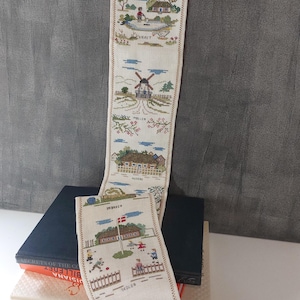 Könnte beinhalten: Eine lange, cremefarbene, bestickte Wandbehänge mit verschiedenen Szenen von Gebäuden, Windmühlen und Menschen. Die Szenen sind in verschiedenen Farben gestickt, darunter Blau, Grün, Rot und Braun. Die Texte "KICKEEN", "MOLLER", "SKOLEN" und "SMEPER" sind in den Stoff gestickt.