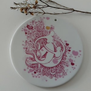 May include: A white ceramic ornament featuring a pink and white design of a mother and child figure surrounded by hearts and swirls.
