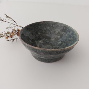 May include: A small, dark green ceramic bowl with a speckled glaze. The bowl is slightly flared at the top and has a rounded bottom.