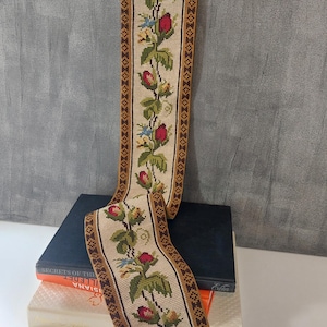 May include: A vintage needlepoint tapestry with a floral design featuring red roses, green leaves, and blue accents. The tapestry is framed with a brown border and has a brass hanging fixture. The tapestry is displayed over a stack of books.