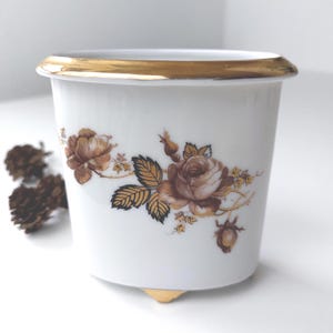 Könnte beinhalten: Weißer Keramiktopf mit goldfarbenem Rand und braunem Blumendesign. Der Topf hat drei goldene Füße.