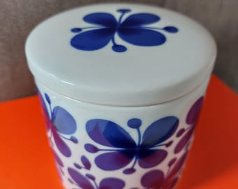 May include: A white ceramic jar with a lid, decorated with a repeating pattern of blue and purple butterfly shapes. The jar is on an orange surface. The lid has a single butterfly design.
