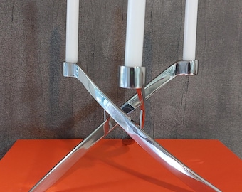 May include: A silver metal candelabra with three white candles. The candelabra has a modern, geometric design with intersecting arms. The candles are white and have a simple, elegant appearance. The candelabra is on top of an orange book.