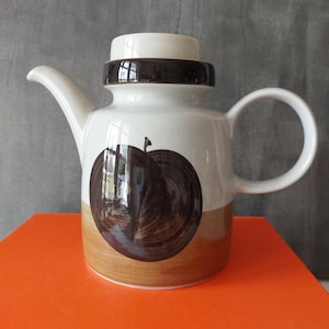 Könnte beinhalten: Vintage Teekanne aus Keramik mit weißem Korpus, braunen Akzenten und einer großen Apfelillustration. Die Teekanne hat einen Ausguss, einen Henkel und einen Deckel. Die untere Hälfte ist hellbraun. Ein klassisches Stück zum Servieren von Tee.