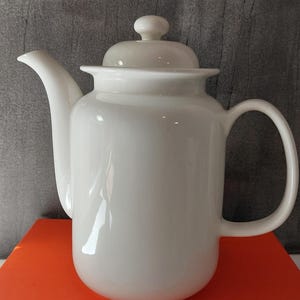 Op de afbeelding: Een glanzend witte keramische theepot met een cilindrisch lichaam, een gebogen tuit, een C-vormig handvat en een bijpassend deksel met een kleine knop. De theepot staat op een oranje boek met de tekst DA LOUISIANA STJAL BILLEDET.