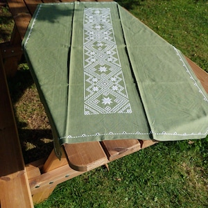 Peut inclure: Un chemin de table vert avec un motif brodé blanc de formes géométriques et d'étoiles. Le chemin de table est posé sur une table en bois.