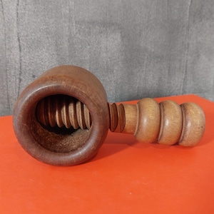 以下が含まれることがあります： 大きな円形の頭とリブ付きの内部を備えた木製のくるみ割り人形。ハンドルは4つの丸みを帯びたセクションで構成されています。 くるみ割り人形は明るい茶色の木材でできており、オレンジ色の表面に置かれています。