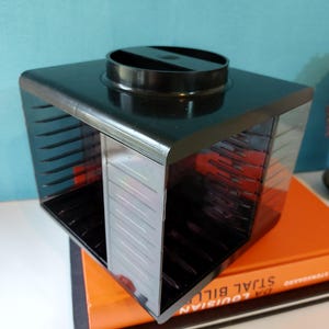 Puede incluir: Una caja de almacenamiento de CD negra y cuadrada con una parte superior circular y una parte delantera transparente. La caja está colocada encima de dos libros con cubiertas naranjas y negras. La caja tiene múltiples ranuras para guardar CD.