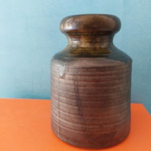 Peut inclure: Un vase en céramique marron avec un corps cylindrique et un dessus arrondi. Le vase a une surface texturée et nervurée et une glaçure marron foncé sur le dessus et le bord. Le vase est posé sur une surface orange avec un fond bleu.